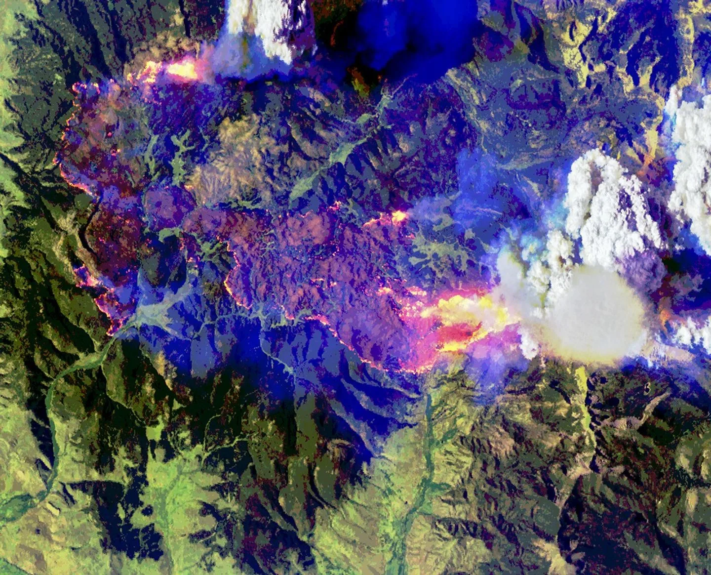 Imagem do incêndio Sand Creek em falsas cores, com pontos rosa e amarelos e nuvens brancas sobre terreno azul e verde.